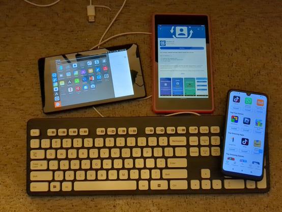 We see two Google Nexus 7 2013 tablets laid out next to each other above a keyboard and phone, all on a carpeted floor. The left tablet is open to the Ubuntu Touch launcher. The right tablet, running LineageOS, is open to F-Droid with the DecSync CC app in F-Droid. On the bottom, we have a gray and white Logitech K310 keyboard. Resting on the keyboard's numpad is a Teracube 2e phone open to the /e/ OS App Store.