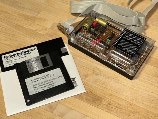 The Fischertechnik Computing interface for PC/XT/AT computers with a grey ribbon cable with a parallel port plug at the end. To the left of it one 5,25" and one 3,5" disk with the driver software.