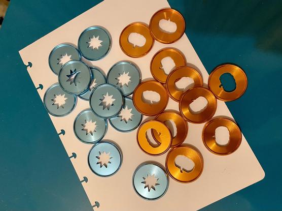 A pile of blue metal discs with a star cut out and orange discs with a pumpkin cutout. They sit on a white A5 bit of paper with 8 mushroom punched holes on its edge, which is on top of a blue surface.