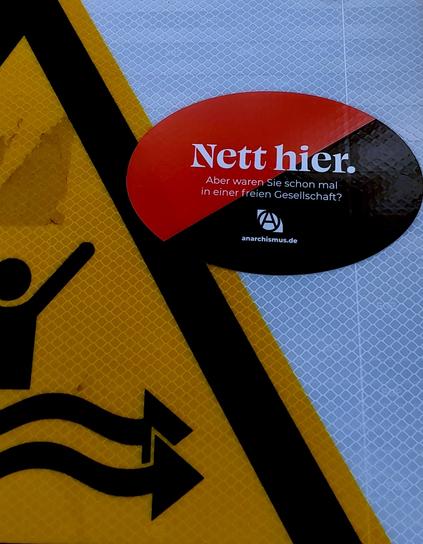 schwarzr und roter aufkleber auf verkehrsschild. auf dem aufjleber steht: "nett hier. aber waren sie schon mal in einer freien Gesellschaft? anarchismus.de"