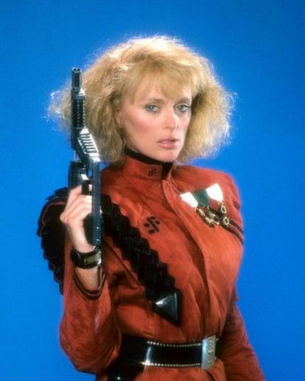In this publicity still, Sybil Danning is standing, wearing the red uniform of the invading aliens.  The red uniform is accented by a thick black belt and strap coming over the shoulder.  She is holding an alien pistol type weapon pointed up beside her head.  Her shoulder length blond hair is frizzy and teased.