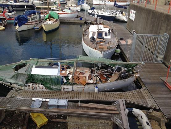 A mastless sailboat with a torn cover revealing a bunch of plywood bulkheads torn out and laying on deck in a messy pile.
