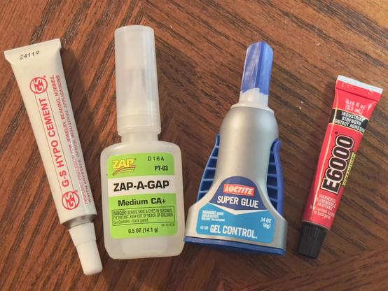 Four varieties of glue for jewelry making lined up on a wood table, from the left: G-S Hypo Cement, ZAP-A-GAP, Loctite Gel Control Super Glue, and E6000 Premium