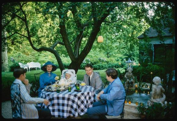 The image depicts a family gathering in an outdoor setting, surrounded by lush greenery and garden ornaments. A group of five individuals is seated around a table adorned with a checkered tablecloth. The adults are engaged in conversation or looking at the camera while one child sits on top of another's lap.

From left to right: Wyatt Cooper dressed in striped pajamas; Chris Stokowski wearing glasses, a coat and tie; Stan Stokowski clad in a suit jacket; Gloria Vanderbilt donning a blue hat, blouse, necklace, pearl earrings, ring, bracelet, watch and holding an umbrella-like object. The child on the table is sitting inside a white highchair with various items like dishes around them.

The photo captures the essence of early 1960s fashion styles and social events in America during that period.