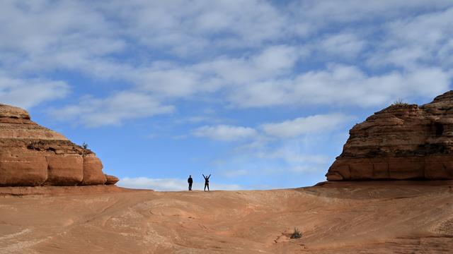 On a saddle of smooth, bare red rock, two hikers stand with a cloud-flecked blue sky behind them.