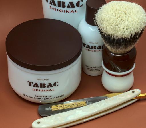 (ChatGPT description)

This image showcases a premium wet shaving setup centered around the iconic Tabac Original line, known for its classic, masculine scent and superior performance.

In the foreground is a large, elegant white tub of Tabac Original shaving soap, marked with its vintage branding and "Since 1959" insignia, highlighting its longstanding reputation in the shaving world. The dark brown lid complements the minimalist design of the packaging, exuding a timeless and sophisticated aesthetic.

Behind the soap tub sits a matching Tabac Original eau de toilette bottle, maintaining the cohesive white-and-brown color scheme. Its compact, rounded design and spray nozzle suggest a convenient and refreshing addition to the post-shave routine, leaving a signature scent.

To the right, a stylish shaving brush with a two-toned handle (cream and rich brown) is prominently displayed. Its dense bristles are perfectly shaped, ideal for whipping up a luxurious lather with the Tabac shaving soap. The craftsmanship of the brush reflects attention to detail and a commitment to quality.

In the foreground, a stunning Bismarck straight razor with a pearl-like handle and a gold-etched blade lies gracefully. Its "Best German Hollow Ground" engraving points to its premium origins and precision engineering, making it a fine tool for a close, traditional shave.