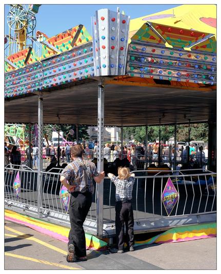 In the image, there is a carnival scene with a variety of attractions and rides, including what appears to be a bumper car ride named "Scooter". Various colorful, lit-up decorative elements can be seen on the ride platform. There are two individuals, likely a father and son, both dressed in similar plaid shirts and jeans. They are standing by the ride's barrier, looking at the activities. In the background, a crowd of people and carnival booths can be seen under a clear blue sky.