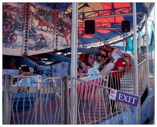 This image depicts an amusement park ride that looks like a spinning or tilting platform ride. The people on the ride appear to be experiencing motion, possibly moving in a circular trajectory based on their postures, leaning back as though reacting to centrifugal force. The decorations are winter-themed, with murals depicting snowy scenes and figures engaging in winter sports such as sledding. The use of vibrant colors in the ride's structure and the canopy contrast with the wintry scenes depicted in the murals. The image further captures a moment of enjoyment and thrill at the amusement park.  The ride operator, a man in red shirt and white cowboy hat, leans on the railing watching the ride with a bored look.   His arm has a thick lettered tattoo reading TORONTO
