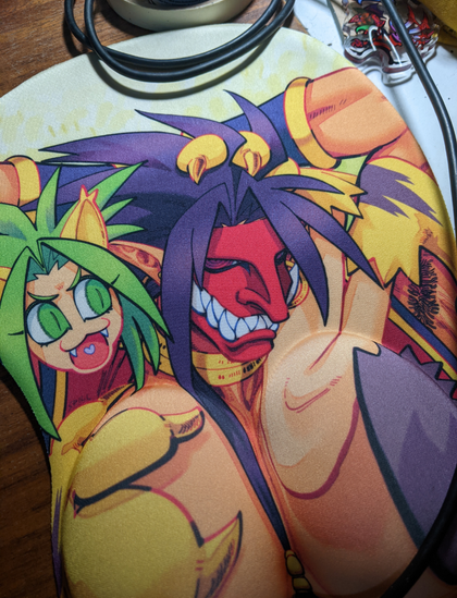 A photograph of a "boob mousepad", a mousepad with an fan illustration of Samurai Shodown's Tam Tam posing with his chest out, the mousepad being filled with round padding where his pectorals are. Cham Cham sits on his shoulders, using her paw to cover one nipple, covering the other with her tip of her tail.