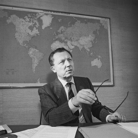 Jacques Delors sitting at his desk with his glasses in his hand. He looks to someone away from the camera. Behind him is a wall-mounted world map.