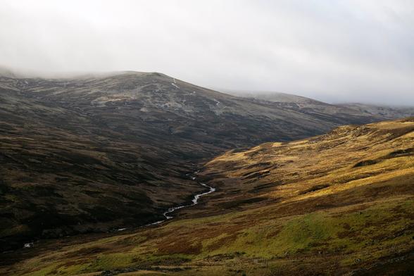 A colour photograph of a rugged landscape. Along the valley a river runs and mist touches the tops of the hills. One side of the valley is illuminated by golden sun.
