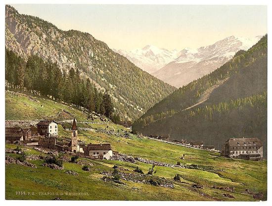 The image depicts a picturesque Alpine landscape featuring quaint village nestled in the valley of an idyllic mountain range. The foreground shows lush green fields with scattered rocks, indicative of rural agriculture or pastures for livestock. Several traditional alpine houses and buildings are spread across the scene, exhibiting wooden architecture typical of this region. Some structures have prominent steep gabled roofs, while others showcase stone walls.

A notable feature in the village is a church tower rising above the other buildings; its spire with a cross suggests it's likely an important landmark for the community. In addition to residential dwellings and farmsteads, there appears to be a larger building resembling a hotel or inn on one side of the valley, suggesting accommodation facilities catering to travelers.

The middle ground is dominated by dense forested slopes leading up to towering mountain peaks in the background. The trees are predominantly coniferous, indicative of high-altitude regions where deciduous species may not survive such harsher conditions at higher altitudes. In contrast, snow-capped mountains peak sharply against a soft-hued sky, hinting that this scene could be captured during late spring or early summer.

The overall ambiance is serene and timeless, evoking the tranquility of rural life within an awe-inspiring natural setting. The image appears to have been professionally printed u [...]