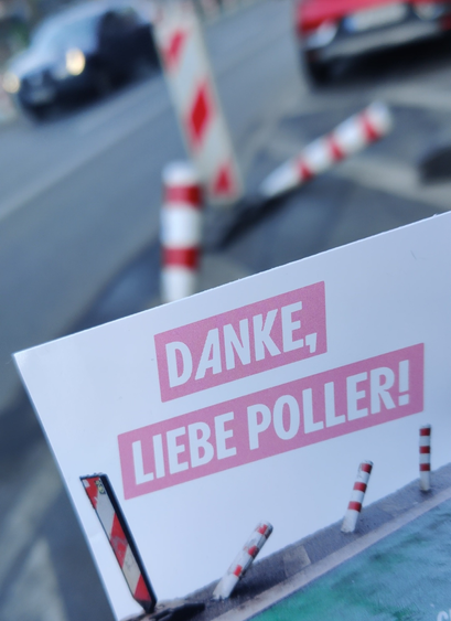 Ein Sticker, auf dem steht "Danke, liebe Poller". Im Hintergrund sind umgefahrene Poller auf einer Straße zu sehen.