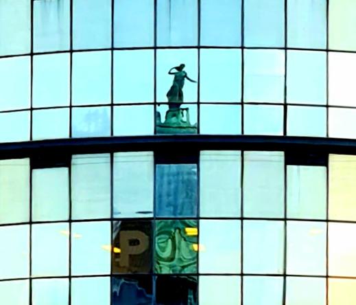 An early 20th Century statue from the top of a public library reflected in a pane of a modern glass-fronted building.