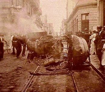 Photograph taken during the “Semana Trágica” in Argentina, in 1919, showing overturned and smoking vehicles, surrounded by crowds. By Unknown author – http://www.upcndigital.org/articulo.php?accID=6381, Public Domain, https://commons.wikimedia.org/w/index.php?curid=4437789