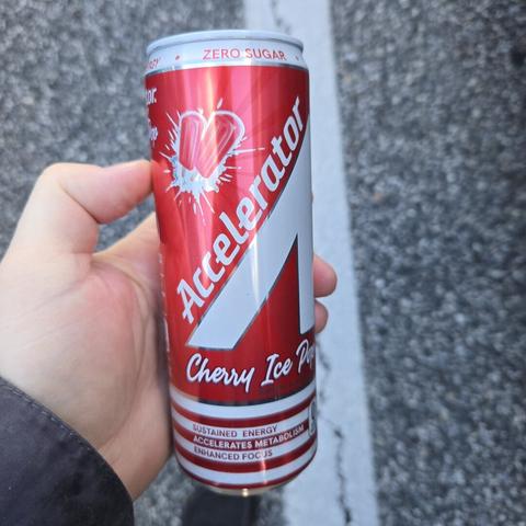 Photograph of a white person's hand holding a red and white can over a concrete ground surface. The can has a stylized white A over a red background with the word "Accelerator" going up the A. There are pictures of a cherry ice pop near the top and it says "Cherry Ice Pop" in white letters beneath the A.