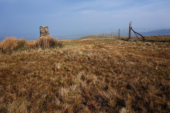 A grassy fell on a cloudy day with a stone built trig point left of centre and a wire fence right of centre.
