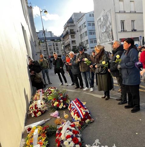 des gerbes de fleurs posées au sol dans une ambiance urbaine. des personnes debout dans une attitude de recueillement, portant des roses jaunes. On reconnaît Danielle Simonnet, Raquel Garrido et Alexis Corbière.