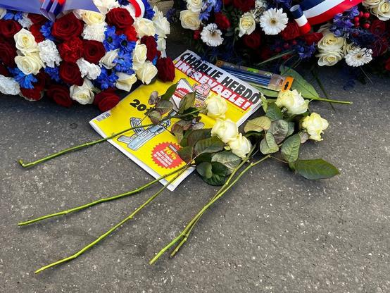des gerbes de fleurs tricolores à l'arrière. à l'avant, des roses jaunes posées sur un numéro de Charlie Hebdo où on peut lire le mot : "increvable !"
