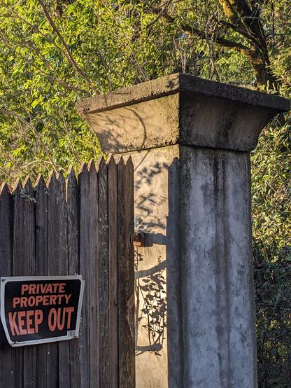 A pillar and a gate. The morning sun casts fine shadows on one face of the pillar, shining through the gap.  On the gate is a "keep out" sign.