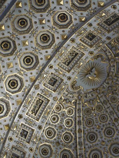 Color photo of a curving, profusely decorated ceiling - executed in white stucco highlighted with gold - that features numerous rectangular and octagonal coffers, each of which contains a rosette at its center