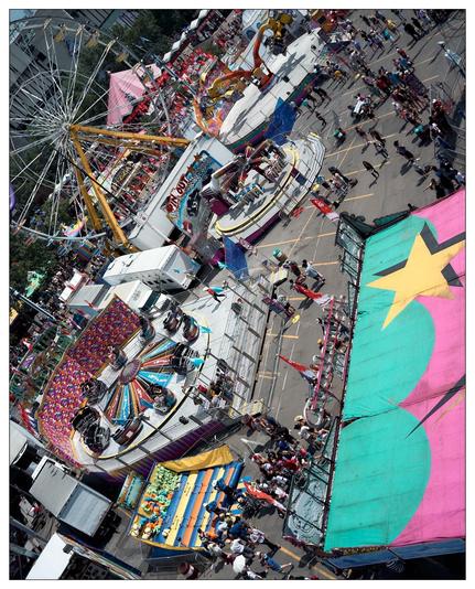 This image captures an aerial view of a bustling fair or amusement park. Prominently featured are various colorful amusement rides including a Ferris wheel, spinning chair swings, and other carnival attractions. Throngs of people can be seen walking around, and there are several game and food stalls line the walkways. The image gives a vibrant, festive feel, highlighted by the bright colors of the attractions and tents.