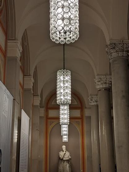 Das Bild zeigt einen langen, hohen Korridor mit hohen Säulen und gewölbten Decken. In der Mitte des Korridors hängt eine auffällige, mehrstöckige Leuchte, die aus vielen kleinen, durchsichtigen, kugelförmigen Elementen besteht.  An den Wänden befinden sich Ausstellungsbanner oder -tafeln. Am Ende des Korridors steht eine Marmorstatue eines sitzenden Mannes. Der Gesamteindruck ist der einer eleganten und historischen Galerie oder eines Museums.