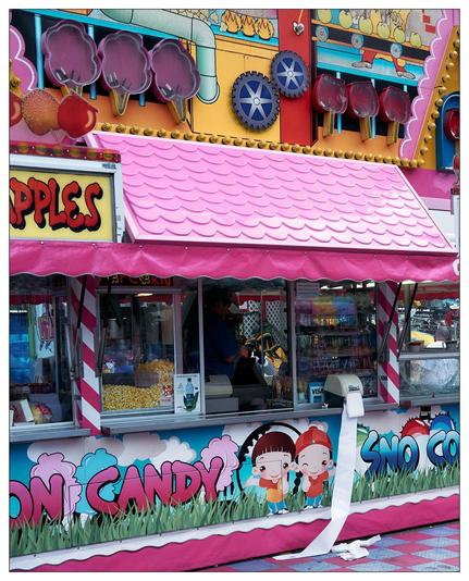 The image shows a colorful concession stand at a fair or carnival. The stand is decorated with bright colors and playful designs including depictions of candies, and a pink roof. It offers various treats such as caramel apples, popcorn, cotton candy, and sno cones. You can see containers of popcorn inside the stand, and there are cartoon depictions of children on the lower part of the cart near the ground. There is also a worker inside the stand, and a point of sale terminal indicating that the stand accepts Visa payments.  A continuous paper towel dispenser has disgorged enough towel that it hangs down to and across the ground.