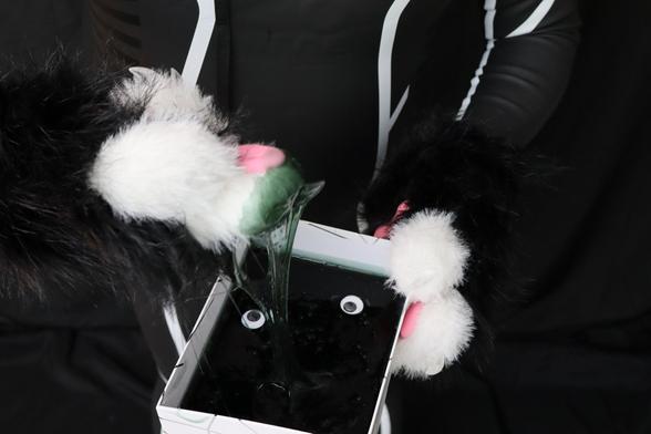 The image shows a person wearing black and white furry paw gloves holding a box filled with a dark, gooey substance that appears to be a type of slime. The slime has two small, round, googly eyes placed on top, giving it a playful or silly appearance. The person is dressed in a sleek, black outfit with white details, resembling a wetsuit or bodysuit. The goo is being stretched out, with some of it hanging from one of the paws.