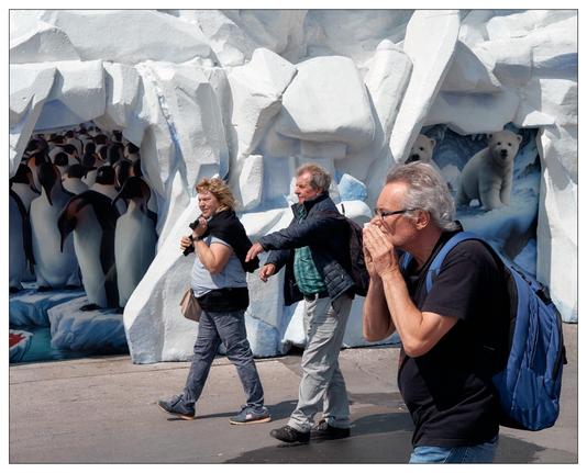 This image features a lively scene with three individuals walking past a large, artificial ice structure that resembles a polar environment. The ice structure is decorated with artistic representations of penguins and polar bears, depicted inside the ice caves. These decorations create a thematic display, possibly part of an exhibit or event aimed at evoking the atmosphere of polar regions. The people in the image, unaware of the camera, are engaged in various actions; one appears to be calling out or covering his mouth, while the others are walking by casually with slight gestures. It’s a colorful, dynamic composition that captures a moment of human interaction against a whimsically designed backdrop.