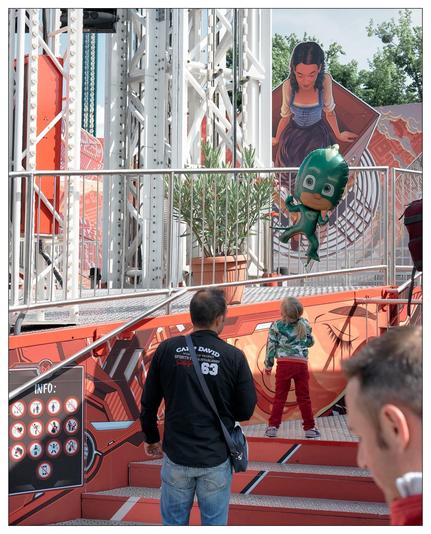 The image features a vibrant and colorful scene, possibly at a theme park or an exhibition. We see a structure with a futuristic design, bright orange and red colors, and metallic elements. Passengers or visitors are navigating stairs and platforms within this structure.

Key elements include:
1. Several people, including two adult men and a child, are interacting with the environment. One man and the child are climbing stairs while the other is looking towards them.
2. Cartoon-style wall murals depicting characters: one appears to be a young girl holding a book with a look of eager curiosity, and the other is an animated green character, which looks like an anthropomorphic duck, floating with a surprised expression.
3. The structure incorporates some greenery with potted plants placed at different levels.

The visual design of the scene is detailed and thematic, likely aiming to create an immersive experience for visitors. The signage indicating "INFO" with icons suggests it's a visitor-friendly space designed to guide and inform guests.