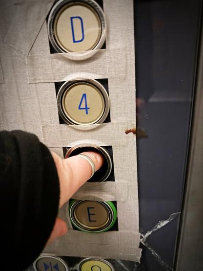Ein Finger wird in das Loch der fehlenden Taste 2 im Fahrstuhl gesteckt, rechts daneben ist ein Spritzer dunkelrote, getrocknete Flüssigkeit