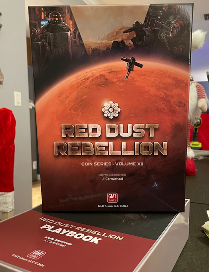 A photo of the box top of Red Dust Rebellion. The artwork on the box has a portion of mars on the cover, with some people in the background with riot shields, others seem to be rioting.