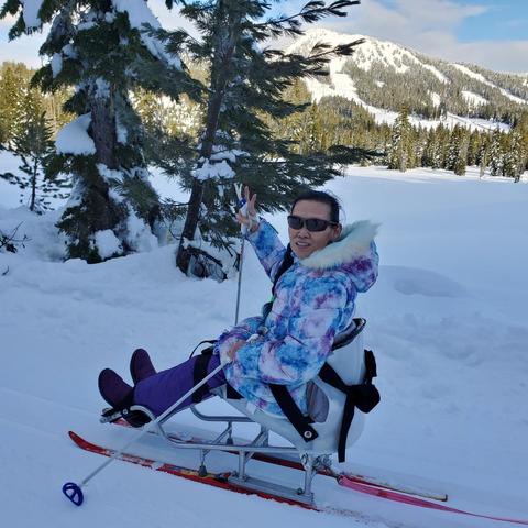Me, wearing sunglasses and a colorful, patterned winter jacket, is sitting in a specialized adaptive ski sled. They are smiling and making a peace sign with one hand while holding onto a ski pole with the other. The snowy landscape features evergreen trees and a snowy mountain in the background, indicating a winter setting.