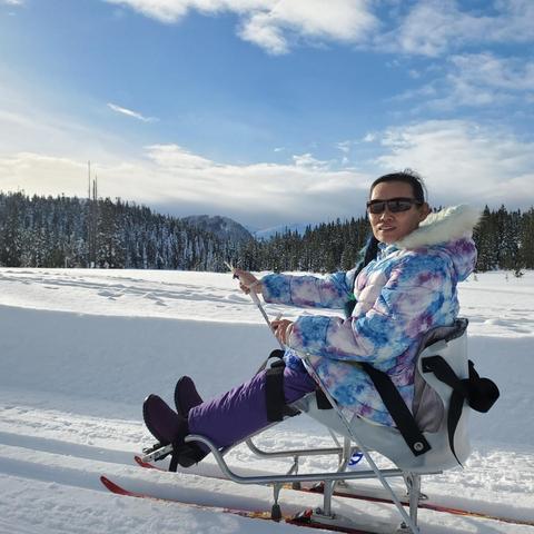 I'm wearing sunglasses and a colorful, patterned winter jacket, sitting in a specialized adaptive ski sled. Smiling. The snowy landscape features evergreen trees and a snowy mountain in the background, indicating a winter setting.
