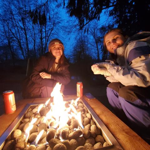 Two women sit by a fire pit filled with stones, smiling at the camera. The fire flickers warmly in the center. One woman wears a beanie and a dark jacket, while the other is dressed in a light jacket and gloves. They are surrounded by a dark, wooded area under a twilight sky, with soft blue light illuminating the scene.