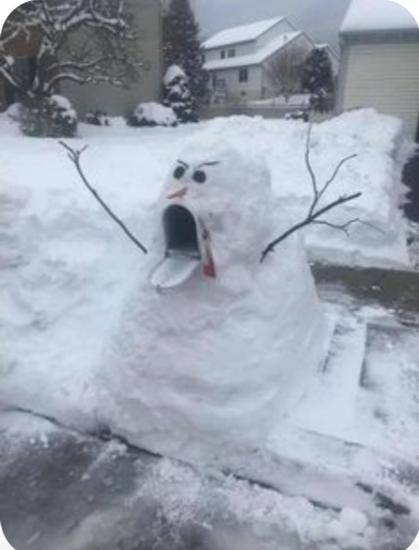 Someone has made a snowman using a mailbox. The snow is stacked around the mailbox, black stones and a carrot have been added to make a slightly frowning face, and the mailbox door has been left open to make it look like the snowman is sticking out his tongue and that his "mouth" is wide open. Sticks have been inserted in the side to look like his arms; they are sticking out and up a bit, as if to startle someone walking past.