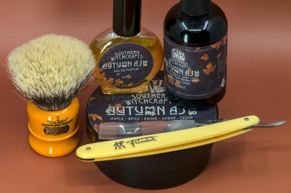 (ChatGPT description)

This image highlights an autumn-inspired wet shaving setup featuring products from the Southern Witchcrafts "Autumn Ash" line, known for its evocative, seasonal scent profile. The arrangement captures the warmth and richness of fall through its design and components.

At the forefront is a Friodur straight razor with a bright yellow handle, standing out against the darker tones of the other items. Known for its precision and durability, this razor is an excellent choice for traditional shaving enthusiasts.

To the left is the iconic Chubby 2 shaving brush with a vibrant orange handle and dense badger bristles. Renowned for its lathering performance, the brush complements the autumnal theme with its warm hue, resembling fall foliage.

The centerpiece of the setup includes a tub of "Autumn Ash" shaving soap, adorned with a seasonal label featuring falling leaves, cobwebs, and moody tones. Its scent, described as a blend of maple, spice, smoke, amber, and cedar, promises a cozy and nostalgic shaving experience.

Behind the soap is a matching "Autumn Ash" aftershave splash in a dark amber bottle, emphasizing the richness and complexity of the scent. Alongside it is the "Autumn Ash" eau de parfum, elegantly bottled, ensuring that the warm and smoky fragrance lingers throughout the day.

This carefully curated setup celebrates the essence of fall, combining functional tools with artisanal products.