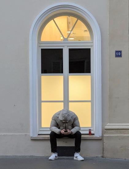 Un  jeune homme assis sur le bord d'une fenêtre dans une rue de Paris. Il est tellement penché sur son téléphone que sa posture pourrait évoquer la tristesse ou l'accablement. La réalité est probablement assez différente.