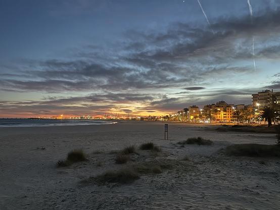 Fotografia d’una platja, a l’esquerra la mar i a la dreta la ciutat. Al fons es veuen els últims rajos de sol grocs després de posar-se, mentre el cel s’enfosqueix i uns núvols grisos el taquen. A la part superior, un estel brillant corona discretament la foto.