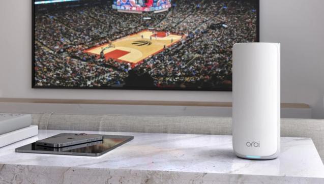 Ein weißer Netgear Orbi 870 steht auf einer Marmorfläche, daneben ein iPad und ein iPhone, im Hintergrund an der Wand ein größerer Flachbildfernseher, auf dem ein Basketballspiel läuft. Foto (c) Netgear.