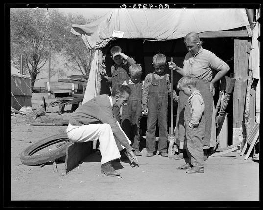 This black and white photograph captures a candid moment between Tom Collins, the manager of a Kern migrant camp during drought conditions in California, and his family. A group of children is gathered around him as he squats down to engage with them on their level. The setting appears to be an outdoor area near what might have been temporary structures for migrants or refugees affected by natural disasters.
The scene includes various objects such as a large tire lying nearby and wooden planks scattered across the ground, suggesting that it may not be an ideal living environment but one where makeshift arrangements are necessary due to displacement. The people in this image appear focused on their interaction with Tom Collins, who seems attentive and interested in conversing or perhaps examining something closely.
In summary, this photograph depicts a poignant snapshot of life for drought refugees during the 1930s California migrant crisis.