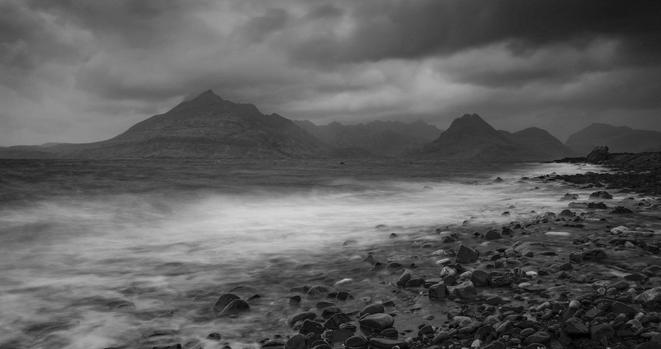 A black and white slow shutter speed photo of the stony beach at Elgol, Isle of Skye on a grey, stormy day with the waves blurred by the slow speed. Cuillin mountain range in the distance.