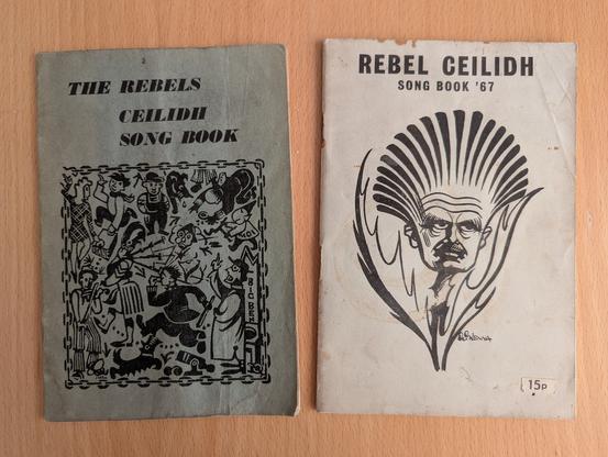 1) Two A5 booklets on a wooden table.
On the left is 'The Rebels Ceilidh Song Book', which has a dark blue-green cover with an ink drawing of lots of people at a raucous ceilidh on it - there are references to political events in Scotland, like the return of the Stone of Destiny.
On the right is the 'Rebel Ceilidh Song Book '67', which has a lighter blue cover with an ink drawing of Hugh MacDiarmid as a thistle. MacDiarmid's face is in the body of the thistle, and its petals closely resemble his famous hair. The thistle's leaves are somehow gesticulating in anger.
Both books are a bit worn.