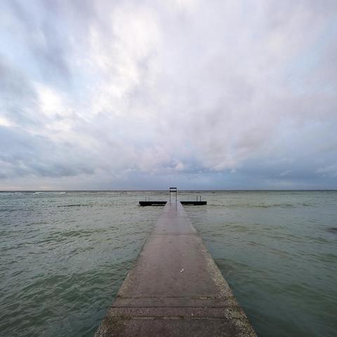 A swimming jetty extends into green sea with rain clouds in the distance.