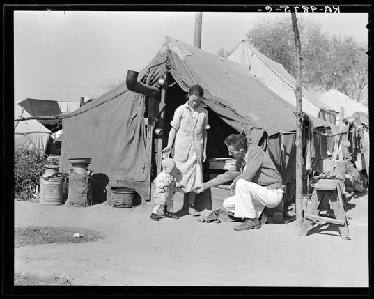 The image depicts a black and white scene from what appears to be the mid-20th century, showing an outdoor setting with makeshift tents. In the foreground, there is a woman standing next to two seated men; one of them seems to be adjusting his footwear while engaging in conversation or offering something to the child he's holding by hand. The tent structure behind suggests a temporary settlement for people who may have been displaced due to drought conditions.

The surrounding area features various objects indicative of daily life and activities, such as buckets, pots, and what looks like cooking implements on makeshift stands. In the background, other tents are visible, hinting at an encampment with several families or groups staying together in close proximity. The overall atmosphere conveys a sense of hardship but also community among those living there.

The caption "Tom Collins, manager of Kern migrant camp, with drought refugee family" suggests that this photograph was taken by Dorothea Lange and is part of her extensive work documenting the lives of migrants during times of crisis such as droughts. The image serves to provide a historical context for understanding how people coped with displacement due to natural disasters like droughts in California's Kern County, which has been an area affected by water scarcity issues over time.

Given this background information and the visual cues within the photograph, one c [...]