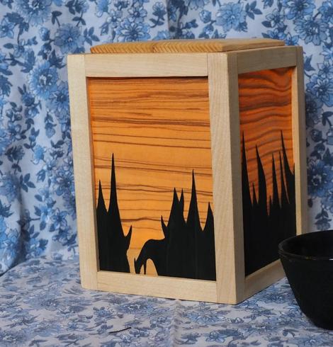 Une veilleuse en bois. C'est un cube et chaque face est faire d'un cadre qui tient une feuille de placage. Le placage très fin laisse passer la lumière depuis l'intérieur de la veilleuse ce qui révèle un veinage et des couleurs intenses. Sur chaque face une deuxième couche de placage empêche la lumière de passer, dessinant une forêt de sapins en négatif.