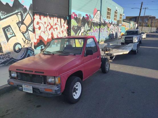 A 3/4 angle shot of an early 1990s red Nissan pickup truck with the bed removed, hitched to a 15 or 20 foot car hauler trailer that dwarfs the size of the little truck. Parked at the curb in front of some impressive colorful graffiti.