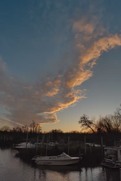 Photo verticale. Dans le quart inférieur, des bateaux amarrés dans le port. En haut le ciel bleu dans lequel flotte une colonne de nuages jaune-orangé. Tout en haut, certains ne sont pas éclairés et sont blanc-gris.