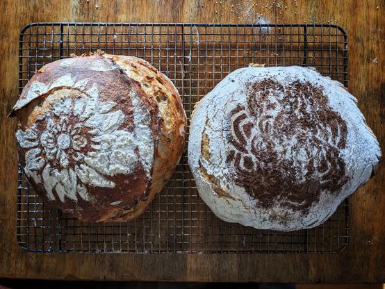 Two loaves of artisan Rustic French bread, baked in the boule style (French for "ball"). One is a golden brown and has an intricate pattern stenciled on top using rice flour, with a full-length score along the entire right side. The other has had rice flour sprinkled all over it, turning the entire crust a silver gray, and a brown pattern has been stenciled on top with cocoa powder. It took has a top-to-bottom score running the entire length of the boule's right side.
